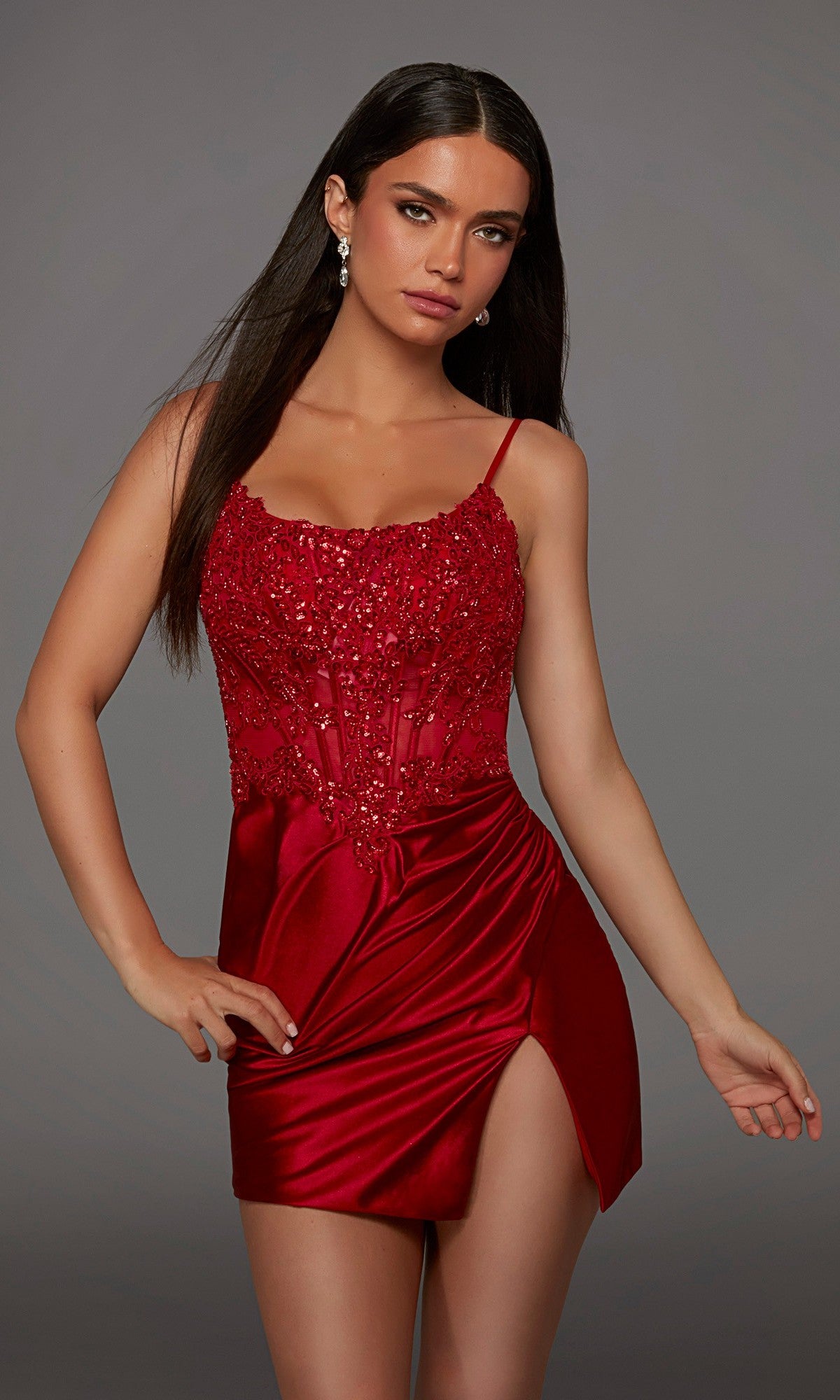 A woman wearing the Short Homecoming Dress: Alyce 40162, a fitted red satin dress with a beaded bodice, spaghetti straps, and a high slit, stands against a plain gray background.